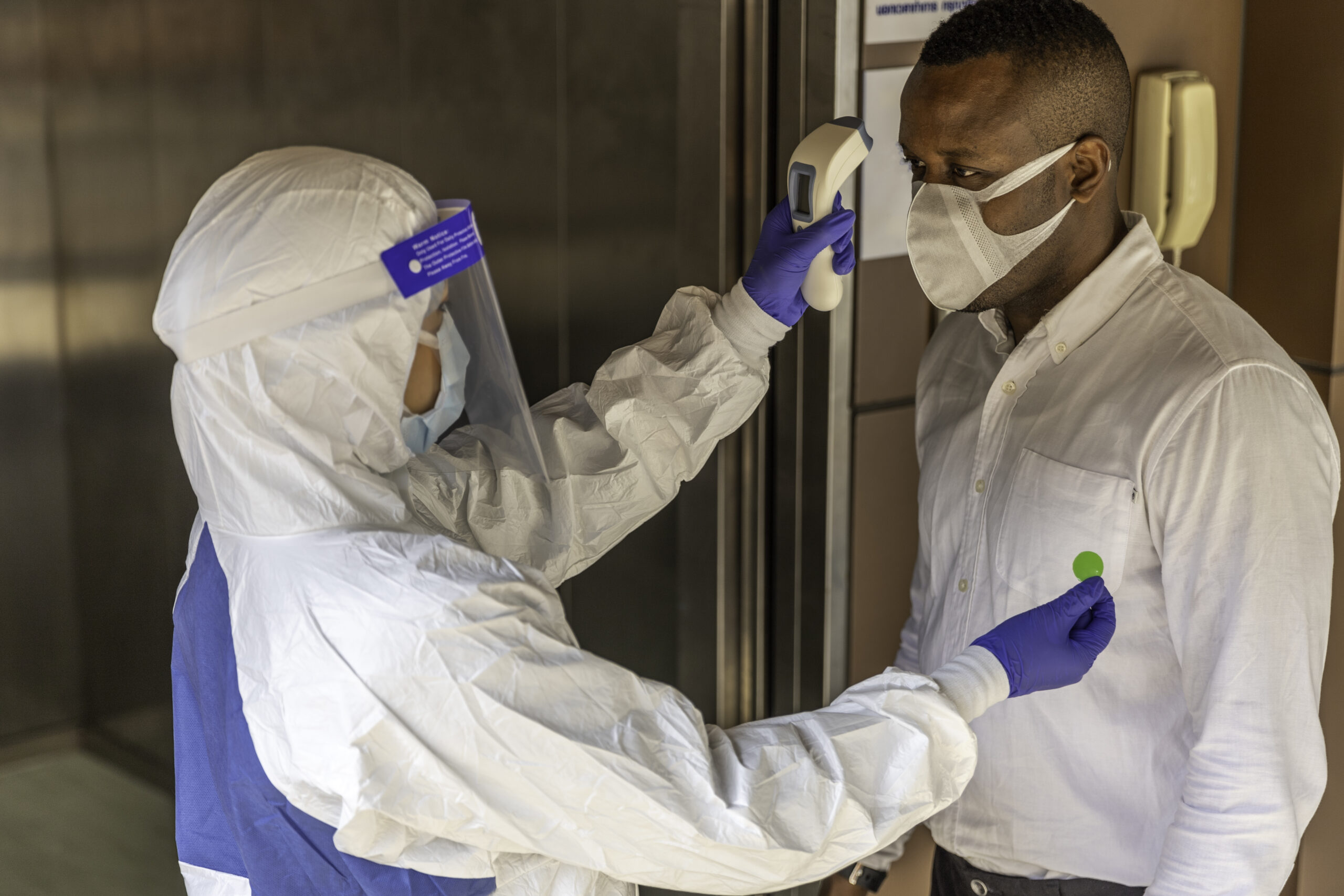 Guard in PPE suit uses infrared thermometer measuring temperature with African male worker scanning for Coronavirus or Covid-19 symptom at office elevator International medical healthcare system